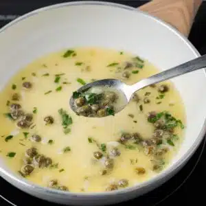 Lemon caper sauce in skillet with a spoon taking some out.