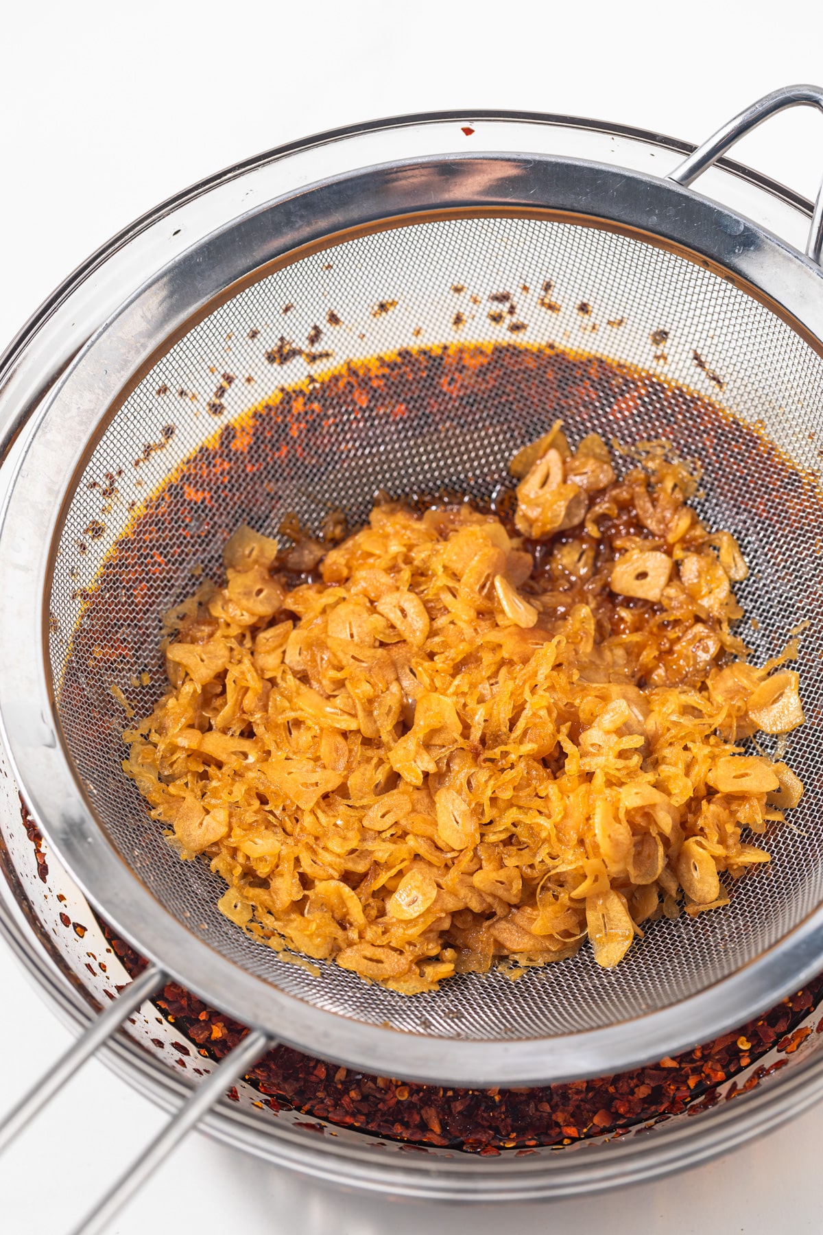 Fried shallots and garlic in a sieve set over a bowl of chili oil.