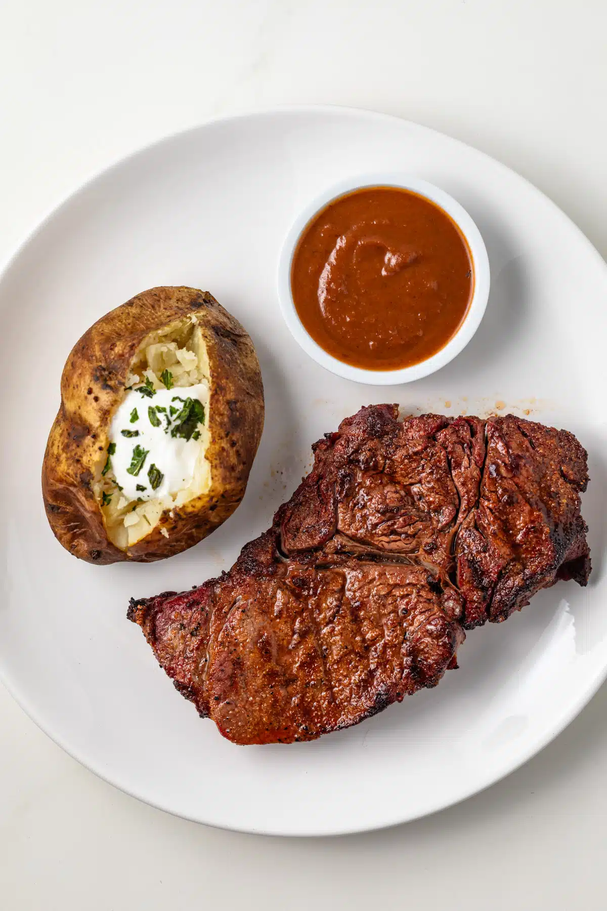 Steak, bake potato, and sauce on a white plate.